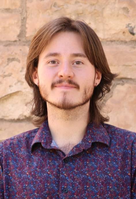 A young man with long hair and a light beard standing in front of a stone wall.