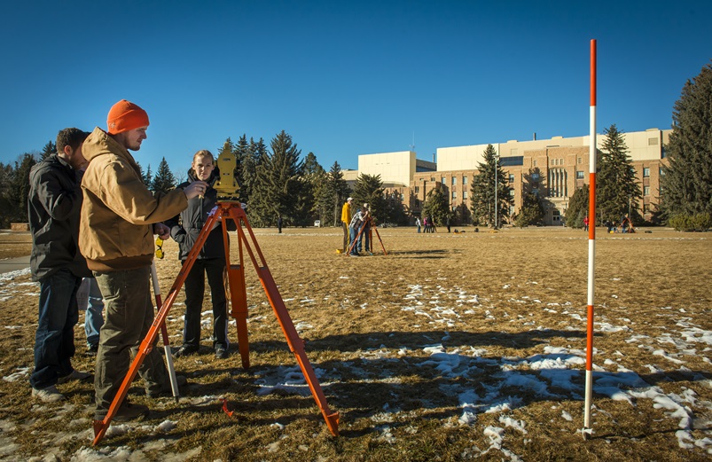 Three students gather around a total station in Prexy's Pasture in late fall. One student is reading the measurements while the other two take notes. More student groups are doing the same in the background