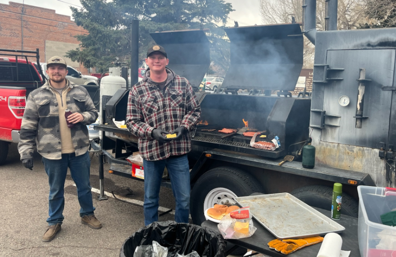 Two students stand in front of a large barbecue, smiling at the camera. The students are grilling food for a student event.