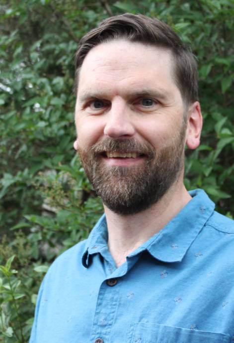 Evan O'Toole, with short brown hair and a beard, wearing a brown shirt, smiles outdoors with blurred trees and rocks in the background.