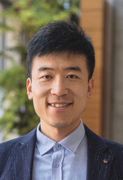 Dr. Pei Li, wearing a light blue button-up shirt and dark blue jacket, stands outside on a sunny day. Blurry trees and part of a building are in the background.