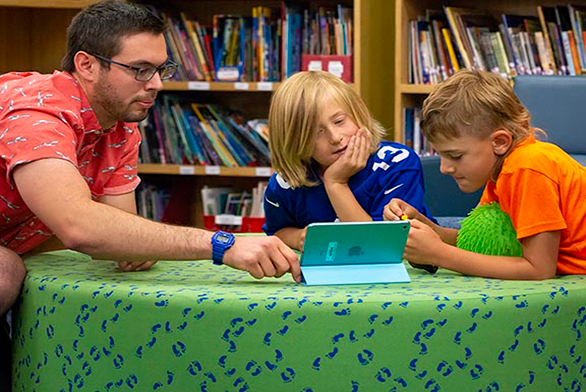 a male tutor reads to two young male students on a tablet device