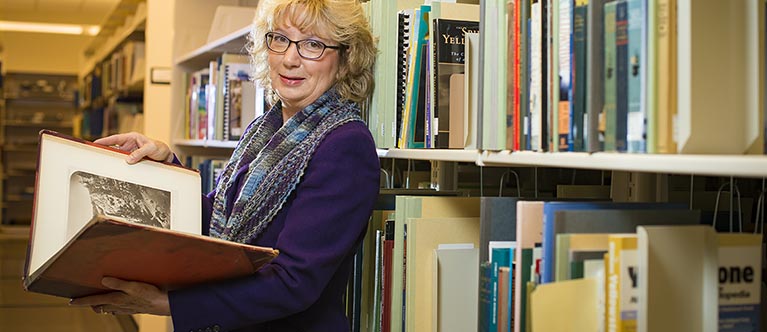 a woman wearing glasses and a purple blazer holds a book while stading in a library