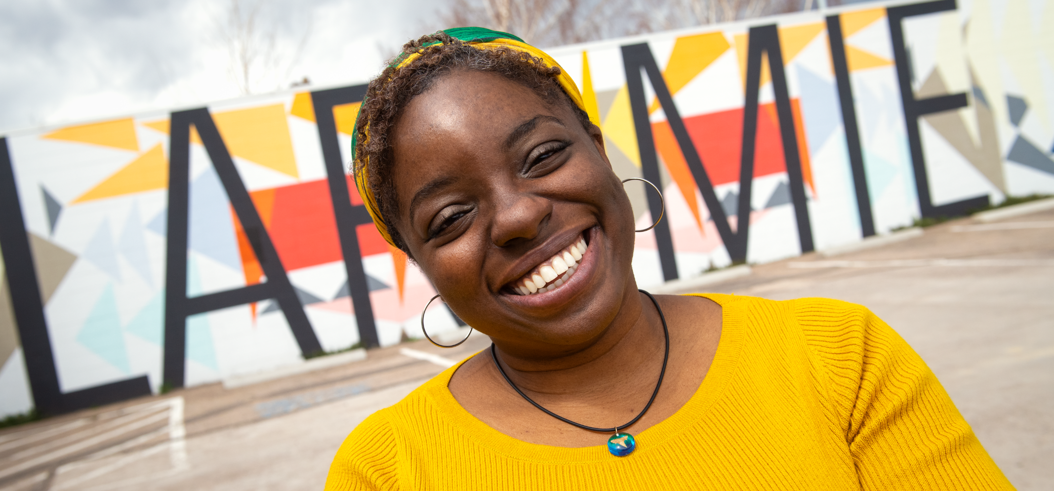 a student in a yellow blouse stands in front of a mural that says "Laramie"