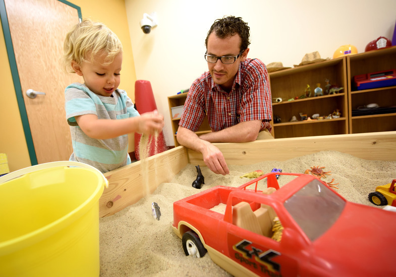Child playing in a sand table with a red toy truck – A young boy pours sand as an adult watches attentively.