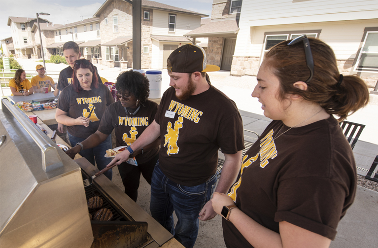 row of five students in brown shirts cooking hamburgers on grill