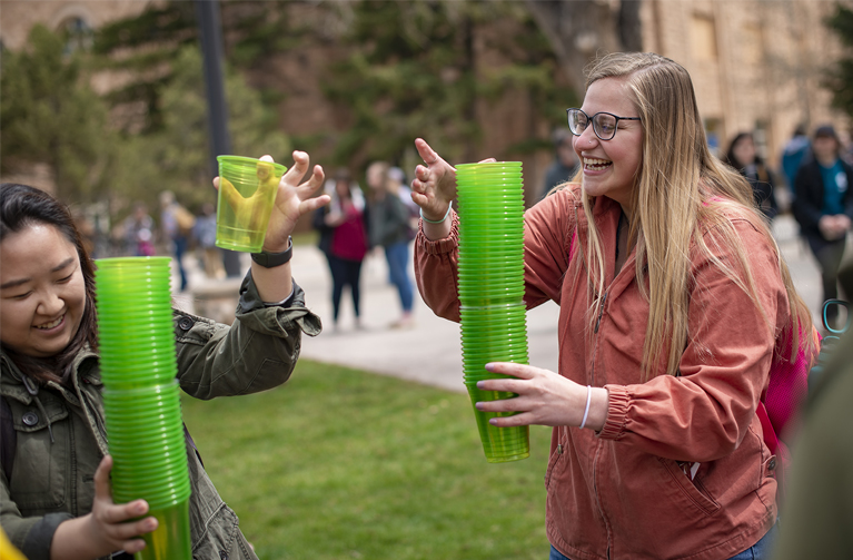 two women laughing and holding stacks of green plastic cups