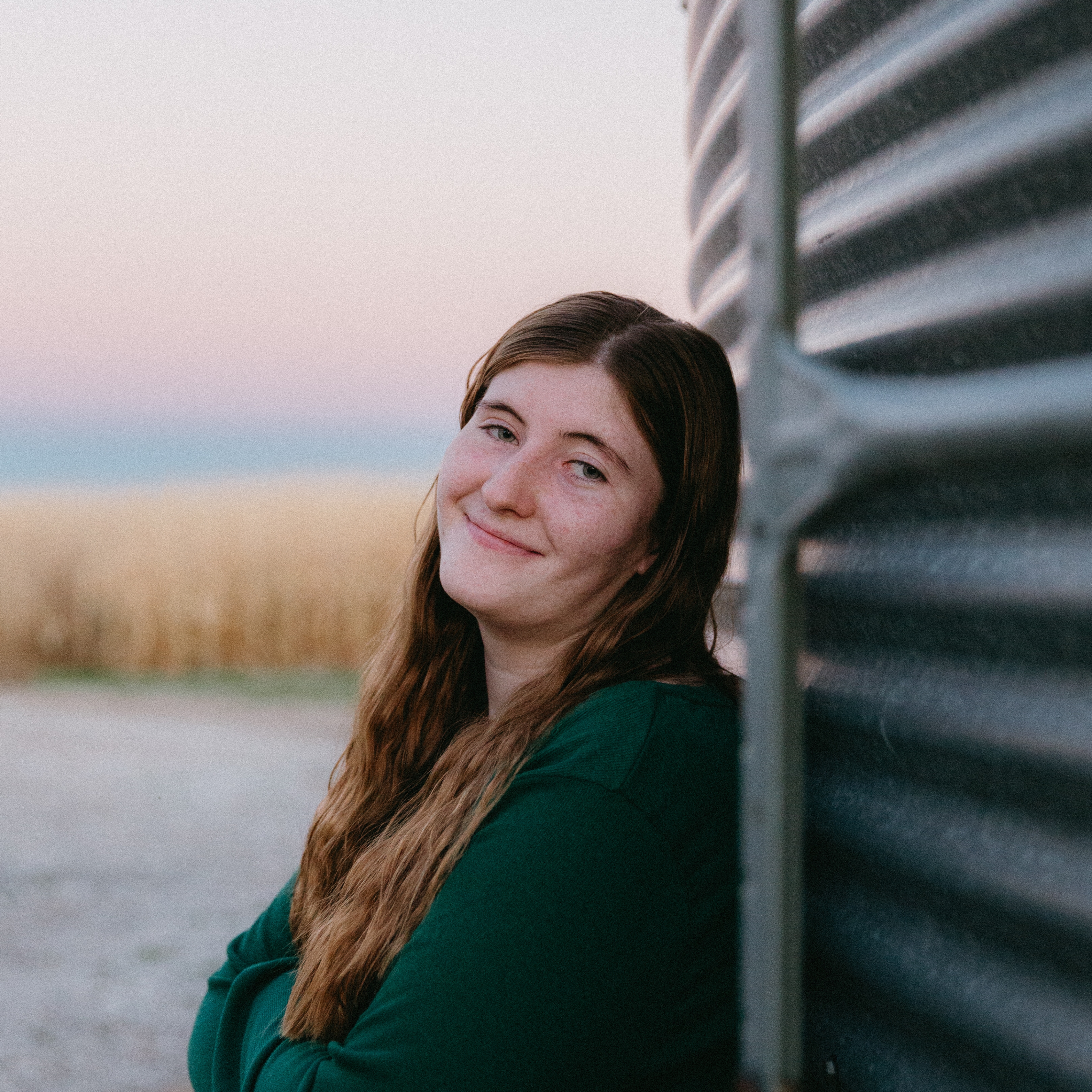 portrait of a woman smiling in front of a corn field and silo. Long brown hair, green long sleeve shirt.