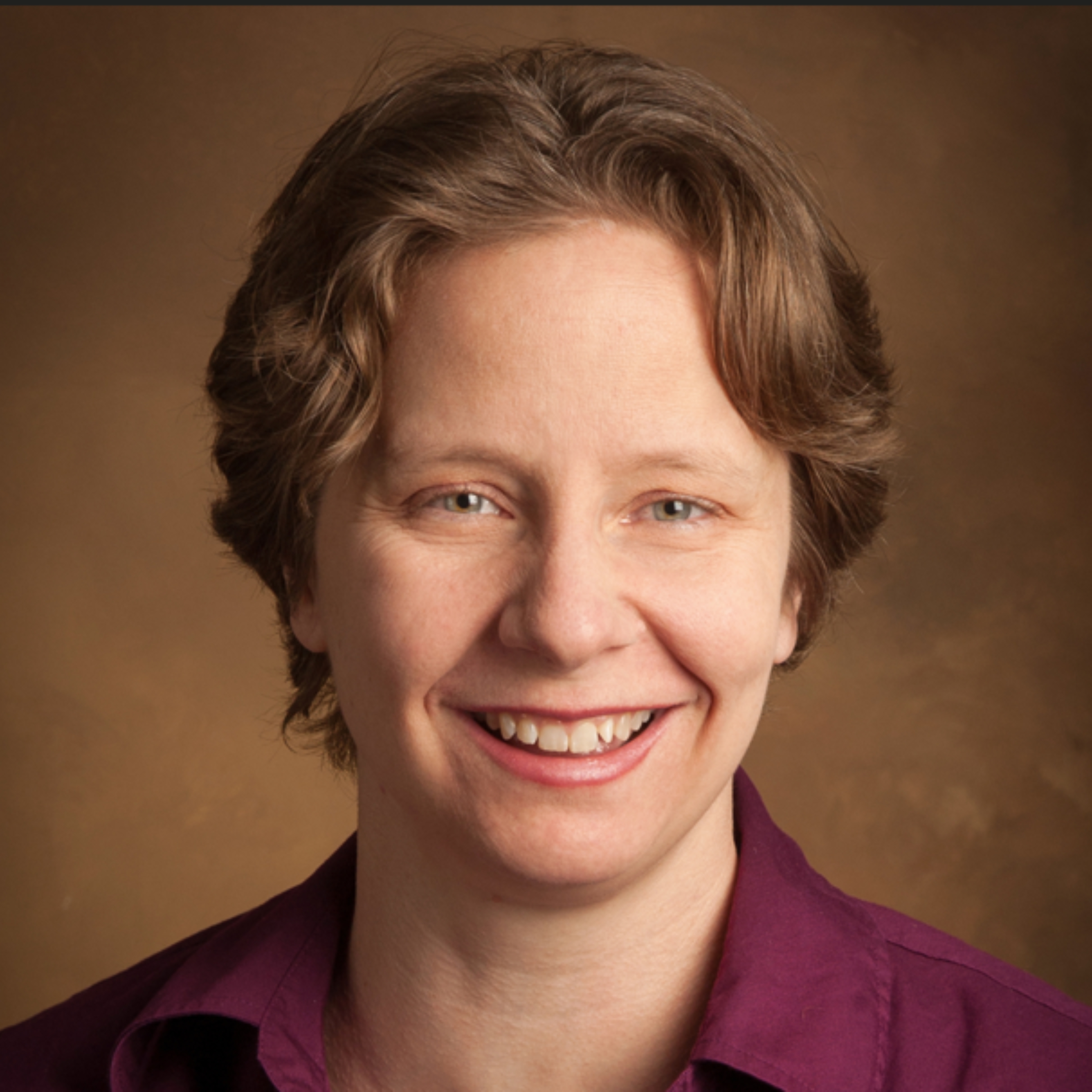 woman smiling in front of a sepia background. brown wavy hair, maroon collared shirt.