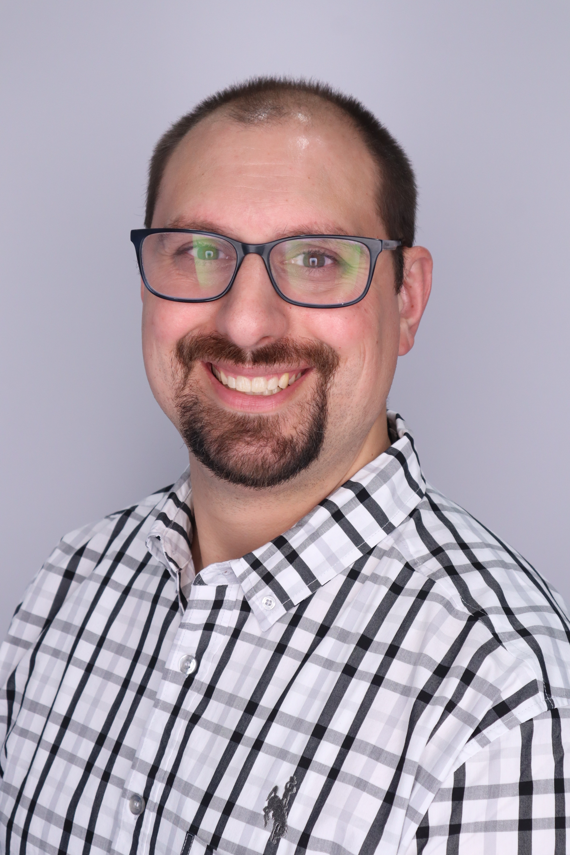 man smiling in front of plain light background. dark hair, dark facial hair, glasses, white and black plaid collared shirt.