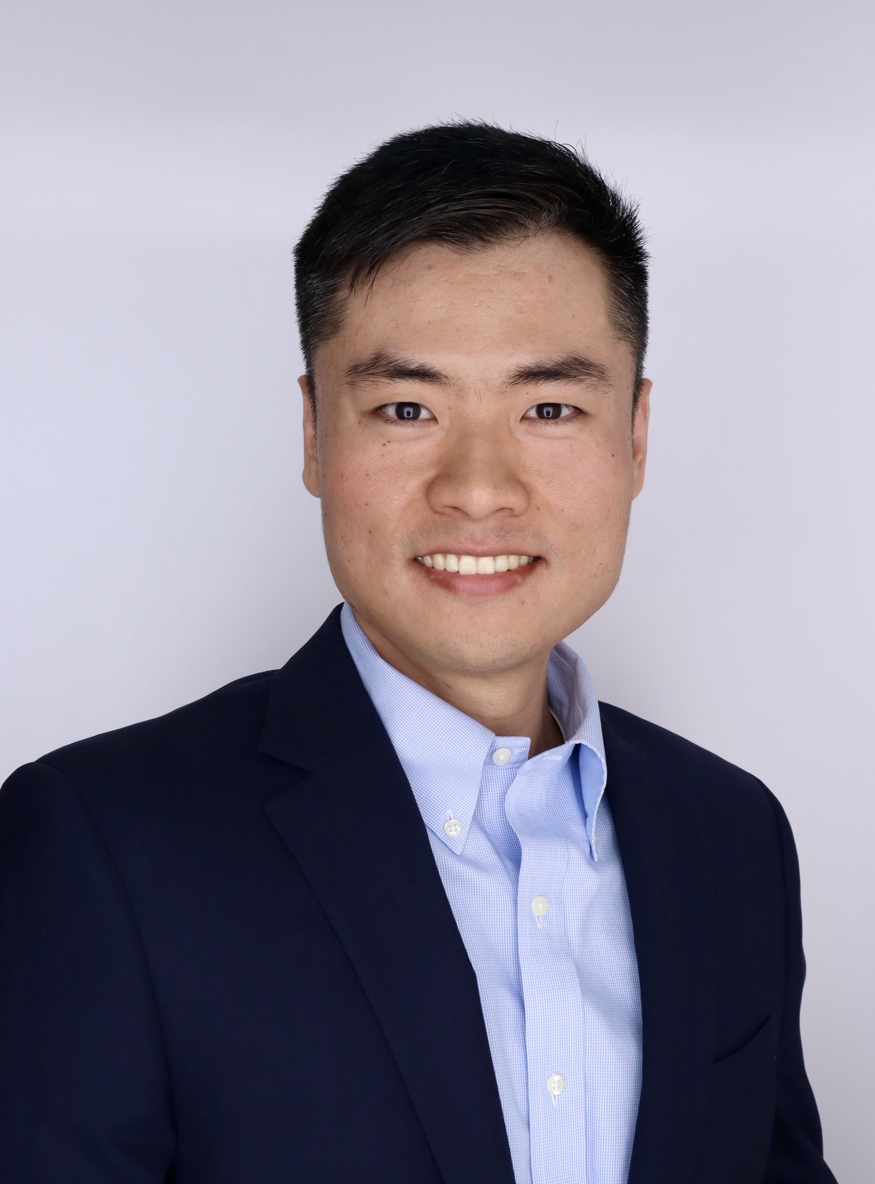 portrait of a man smiling in front of a plain light background. dark blazer with a light collared shirt underneath.