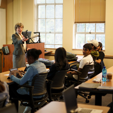 A professor stands at a podium, gesturing towards seated students in a classroom with large windows. Several students face the professor, taking notes or looking at their devices. 