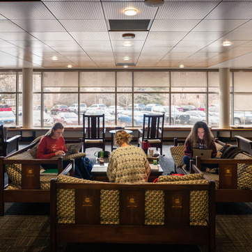 Three people sit on couches in Coe Library, working on laptops and papers, with large windows showing parked cars and trees outside.
