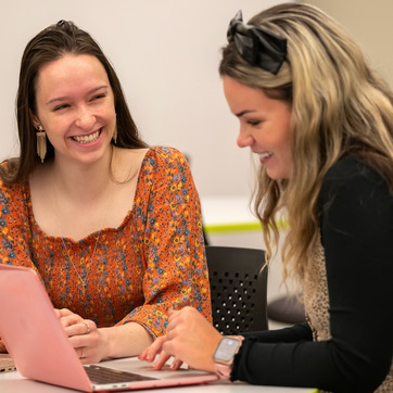Two young women sit together at a table, smiling and looking at a pink laptop. One is wearing an orange patterned top, and the other has a headband and a smartwatch. They appear to be collaborating or studying. 