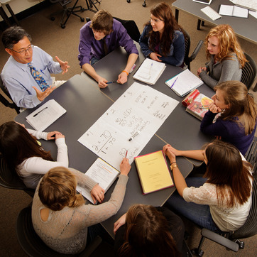 A teacher and seven students sit around a table discussing diagrams and notes on a large sheet of paper in a classroom setting.
