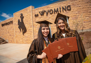 Two UW graduates stand in caps and gowns with their degrees, in front of a University of Wyoming building.
