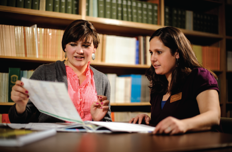 A student meeting with a staff member to review paperwork in a room with shelves of books behind their table