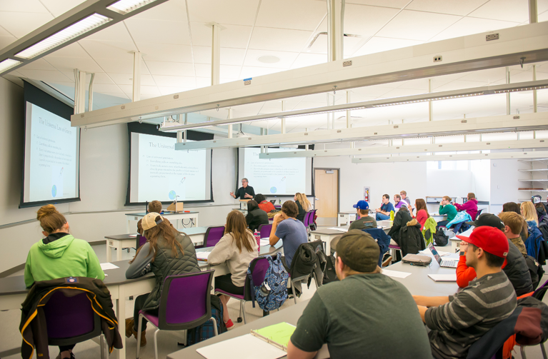 Students sit in a classroom and look at the instructor that is giving a lecture in front of three video screens