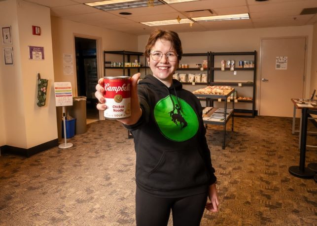 Food Security Coordinator holds up a can of soup in front of shelves in the Pokes Pantry