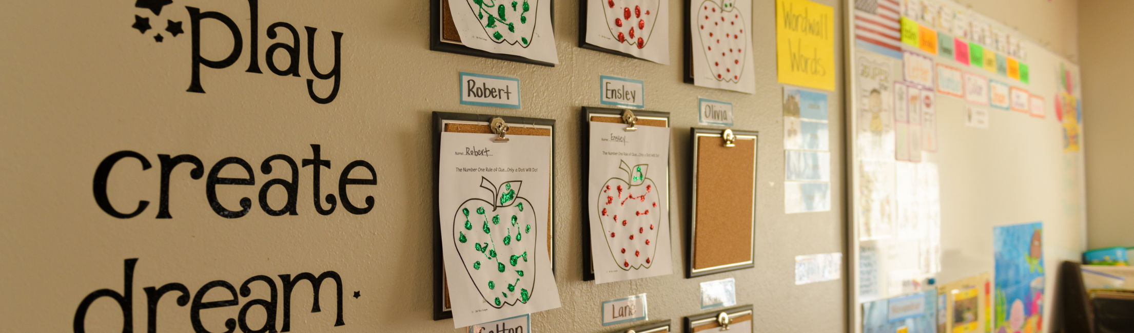 Classroom wall with children's artwork displayed on clipboards, alongside the words 'play, create, dream' in decorative lettering.