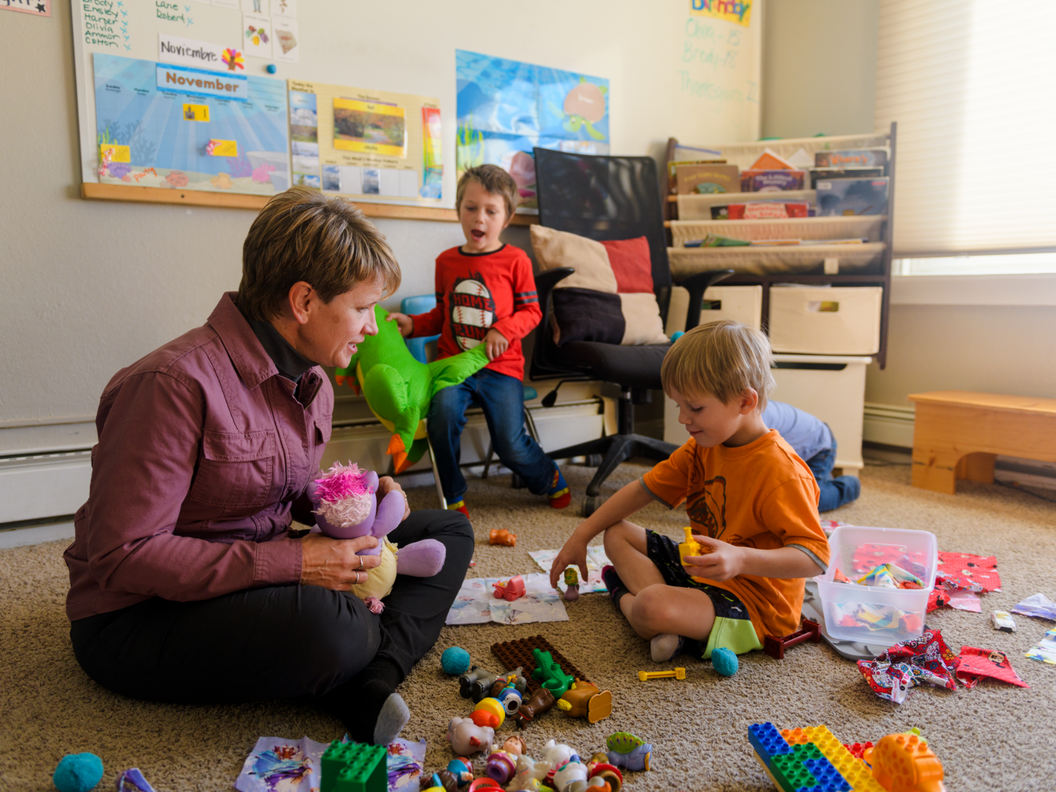 A teacher sitting on the floor engaging with young children in a classroom, surrounded by toys and learning materials