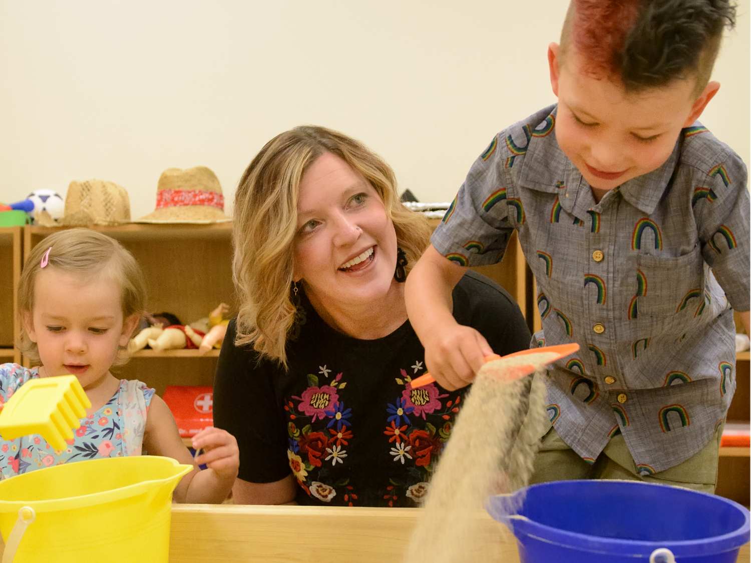 A teacher smiling while engaging with two young children playing with sand and buckets in a classroom setting