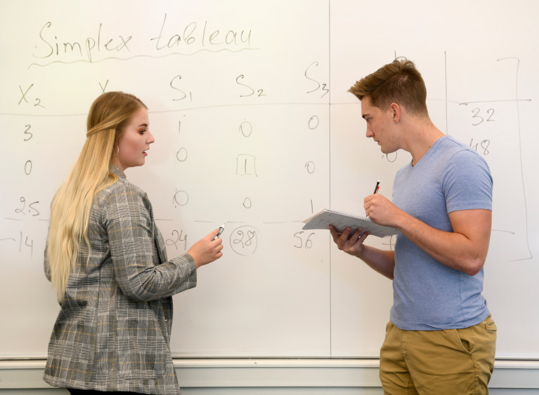 two students solving a math equation on a white board