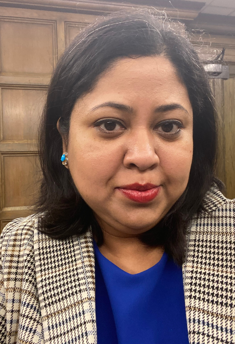 Debolina Glaser in a blue blouse with a tan and black tweed blazer in front of a wood panel background.