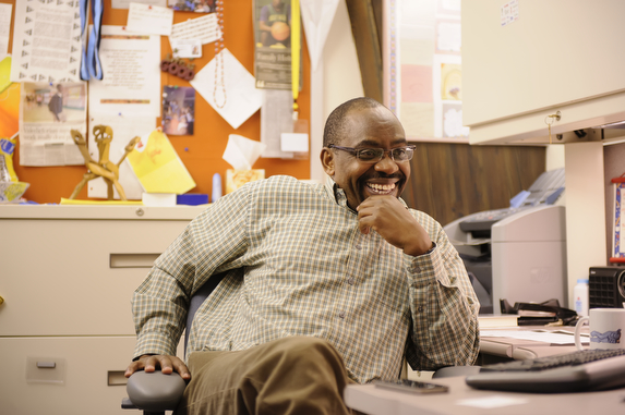 an african american man sits at a desk in a faculty office smiling