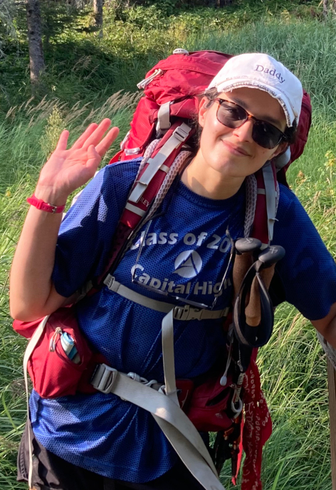 Woman wearing a red backpack waves at the camera.