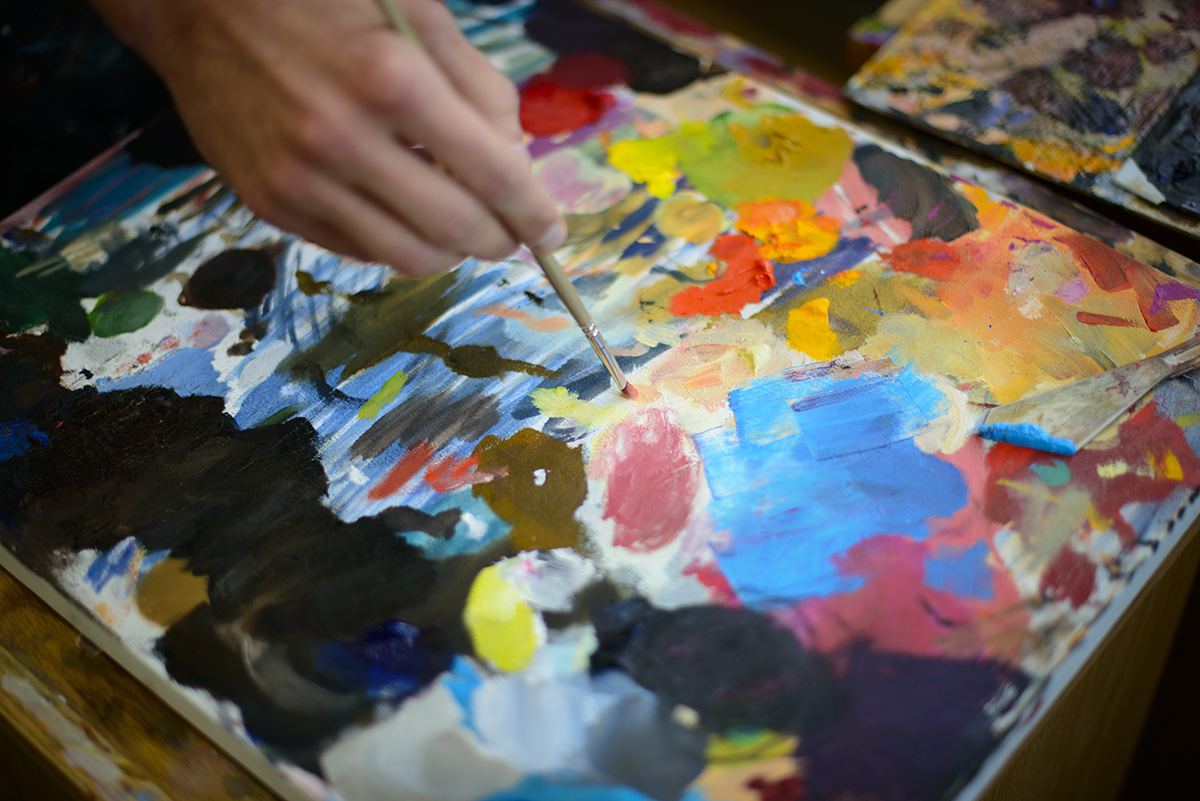 artist hand holding paint above pallet of paint