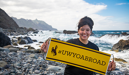 student with #UWYOABROAD sign poses on shore of another country