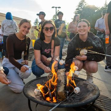 Three students roast marshmallows over an open fire.