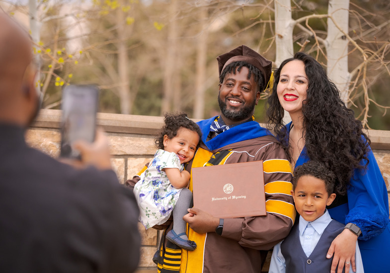 A UW graduate poses with their family and degree in front of a University of Wyoming sign.