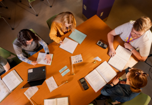 overhead photo of a table scattered with textbooks and papers with students sitting around studying