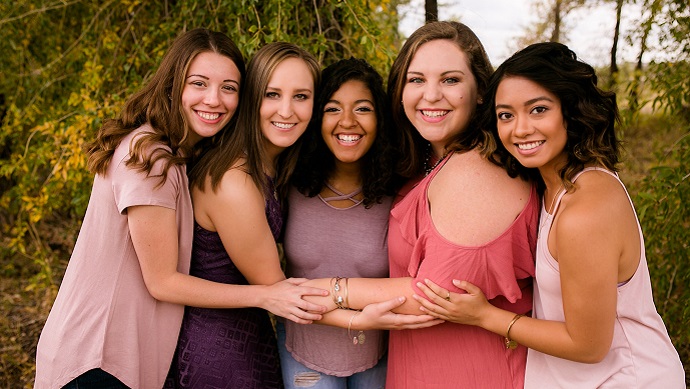 Five girls posing for a group photo in jeans and light purple and pink shirts