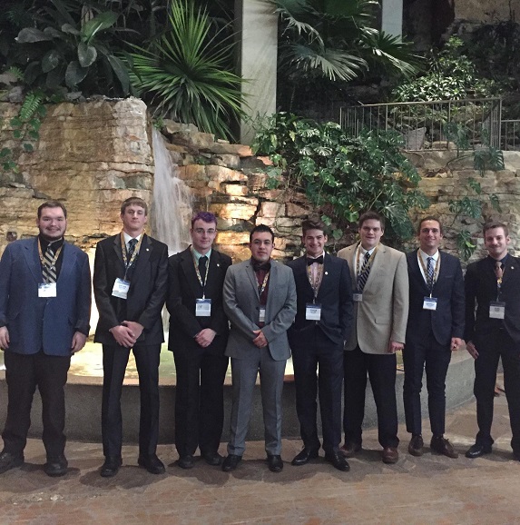 Eight men, all in suits posing for a photo at their fraternity's national conference