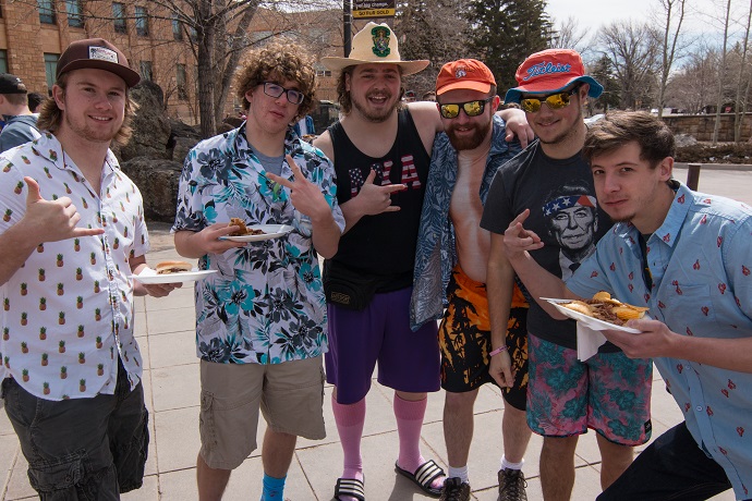 Six members in Hawaiian shirts and various hats holding food  from a bbq