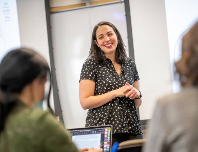 A teacher smiles in the front of the classroom, two blurry students are in the foreground.
