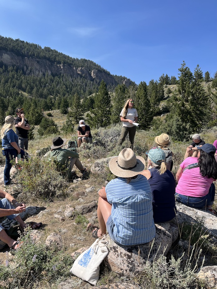 A group of people sit outside on a hill listening to a speaker