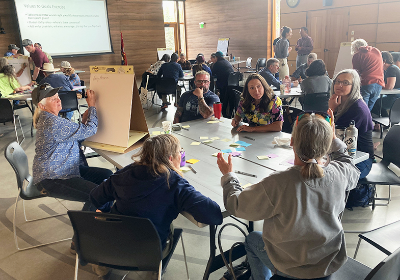 Groups of people sit around tables having small group discussions. In the foreground, the people gesture while one person records their thoughts on a flip chart.