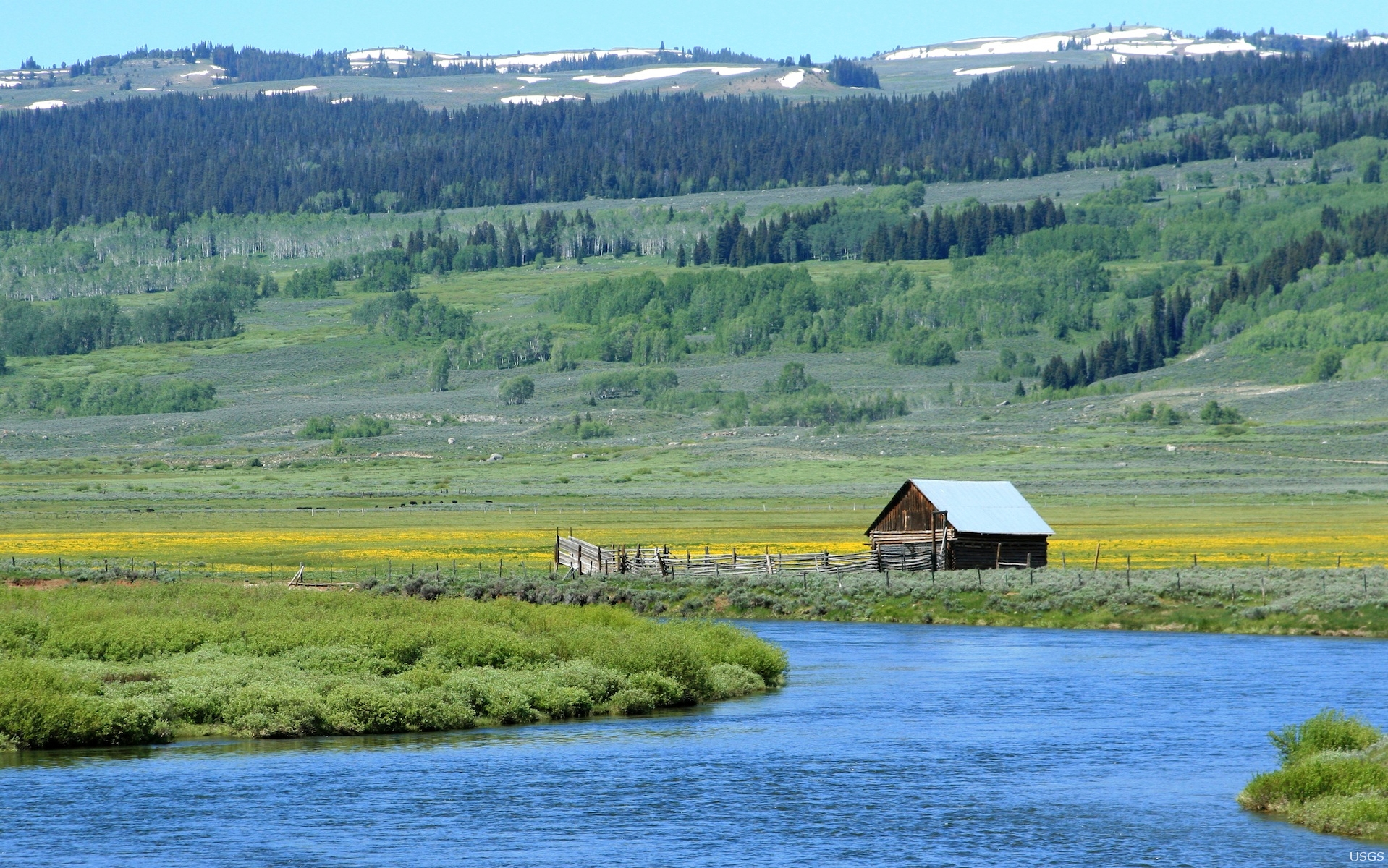 Picture of a cabin along a blue river with snowy foothills in the background