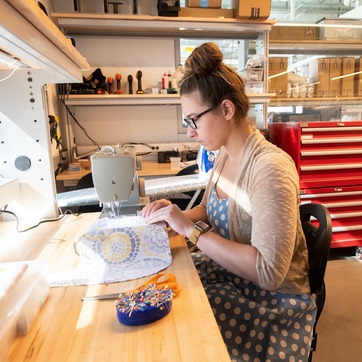 Student sitting at a wooden bench with a sewing machine working on sewing fabric.