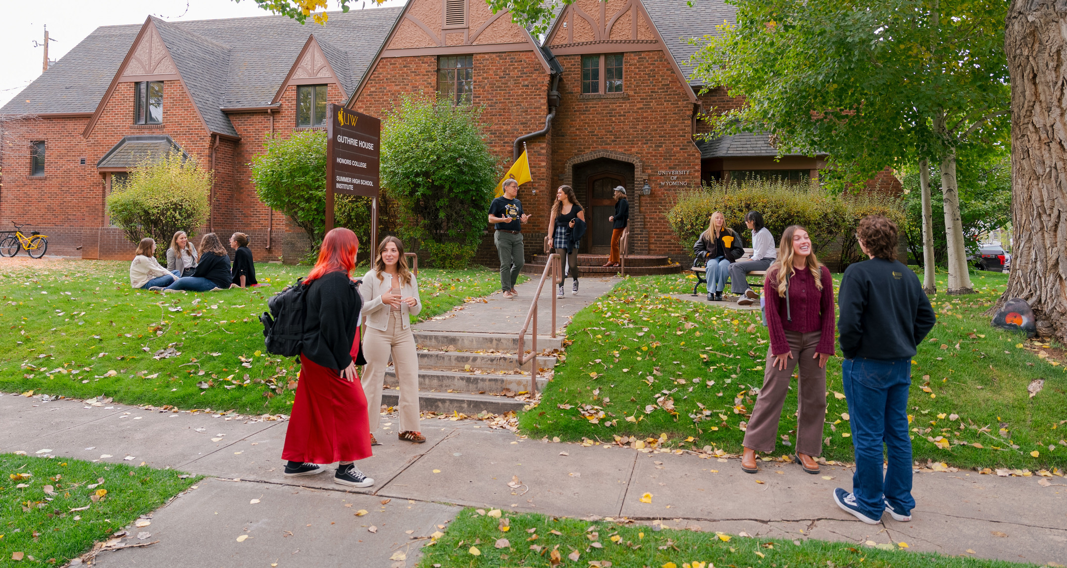 Many different students walk and sit conversing in front of a red brick building.