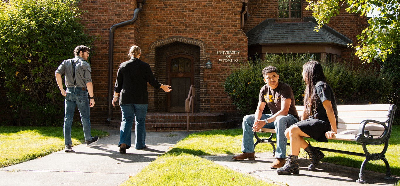 A red-brick, historic house called the Guthrie House featuring a front lawn and trees with students sitting on a bench and two people walking away from the front door.