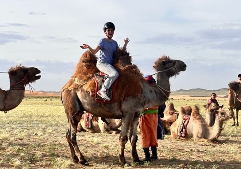 Hallie Cozzens sitting on top of a camel in a yellow grass landscape surrounded by other individuals and camels.