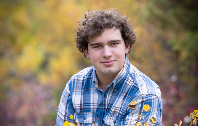 Wolfgang Wuerker wearing a blue and orange plaid shirt in front of a blurred natural background with yellow foliage surrounding him.