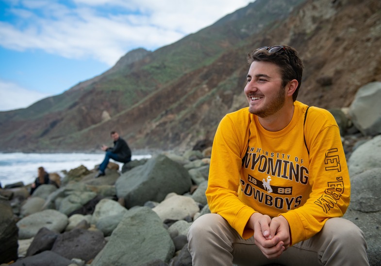 Student sitting on a rocky, beach landscape looking towards his right shoulder. 