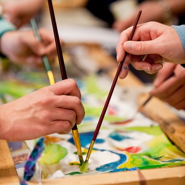 Several hands hold paintbrushes, collaboratively painting a colorful abstract design on a flat surface.