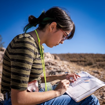 A person sits on a rocky outcrop, sketching in a notebook under a clear blue sky, immersed in nature.
